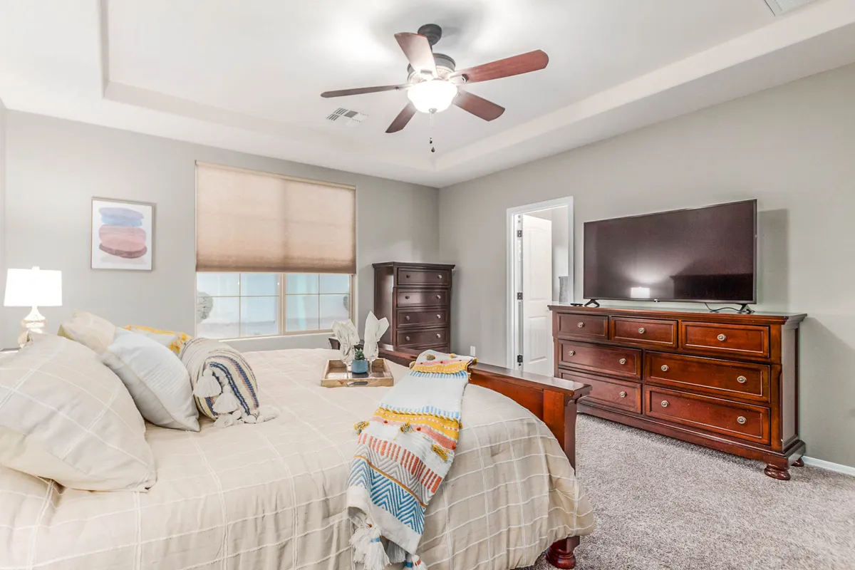 This image features a bedroom with a neatly made bed adorned with a decorative throw, a wooden dresser with a television on top, and artwork on the wall. Natural light filters in, creating a calm and peaceful atmosphere ideal for rest. The presence of a TV and dresser emphasizes comfort and functionality.