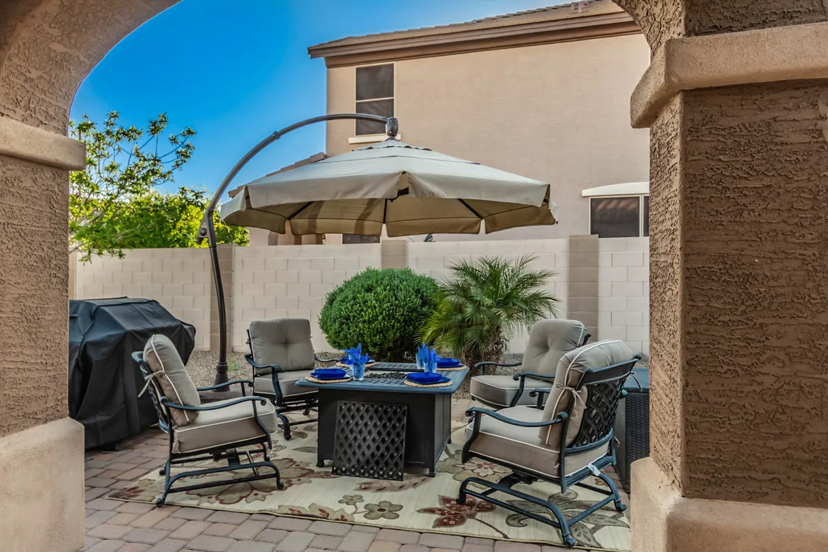 A tranquil outdoor seating area with comfortable chairs, an umbrella for shade, a coffee table, and abundant greenery. The setup is ideal for dining outdoors, socializing, or casual relaxation, and hints at the presence of a firepit with cozy evening ambiance.