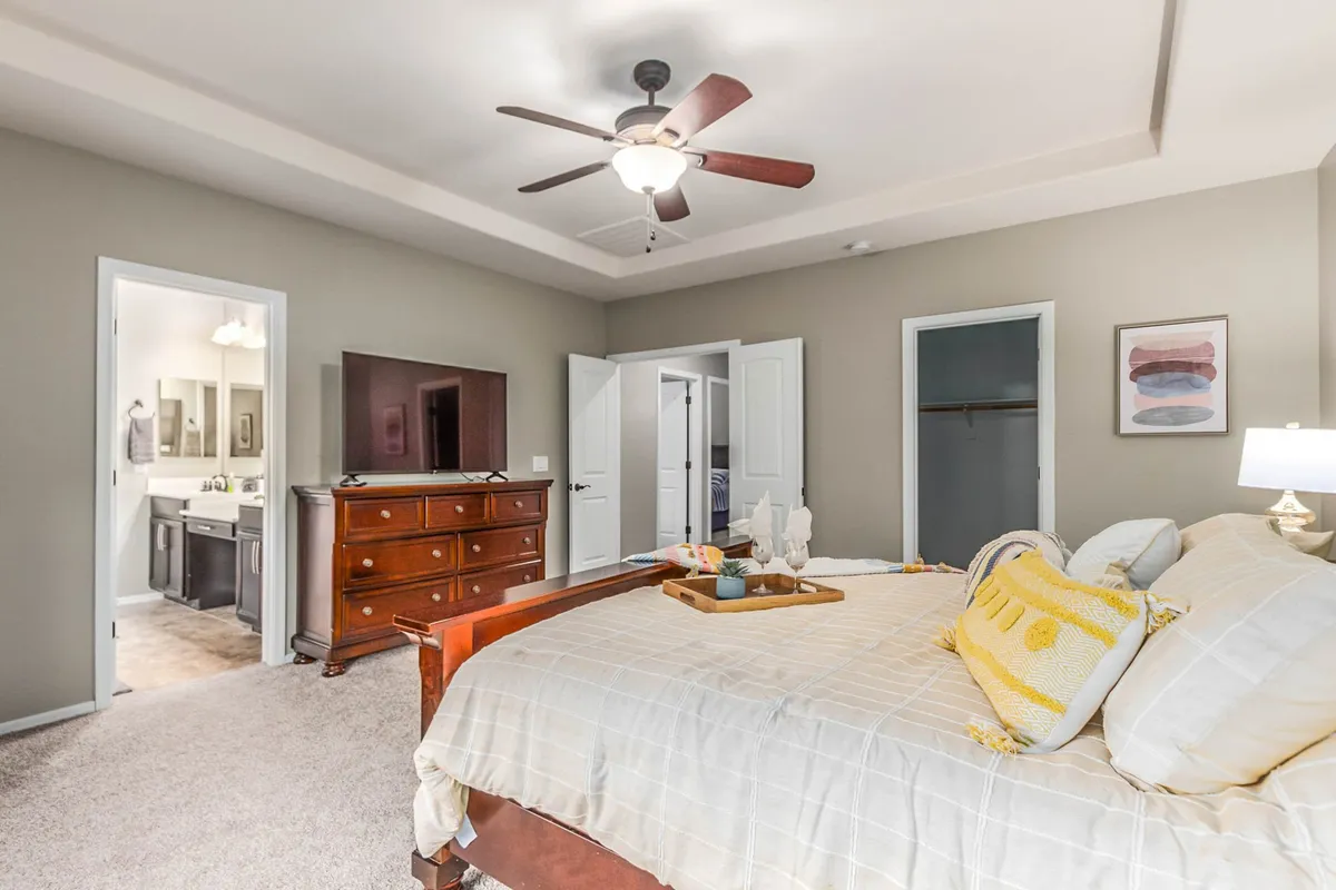 A tranquil bedroom with a bed dressed in decorative pillows, a wooden dresser topped with a TV, open closet doors, and framed artwork. The room is bathed in soft light, emphasizing the feeling of comfort, space, and personal retreat.