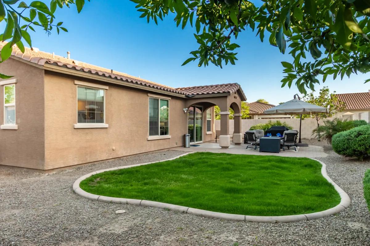 This image displays a green lawn bordered by a patio area with a shaded seating arrangement. The arched entrance and lush garden landscaping add to the welcoming, tranquil atmosphere, and the patio seating provides a dedicated spot for outdoor dining and relaxation.