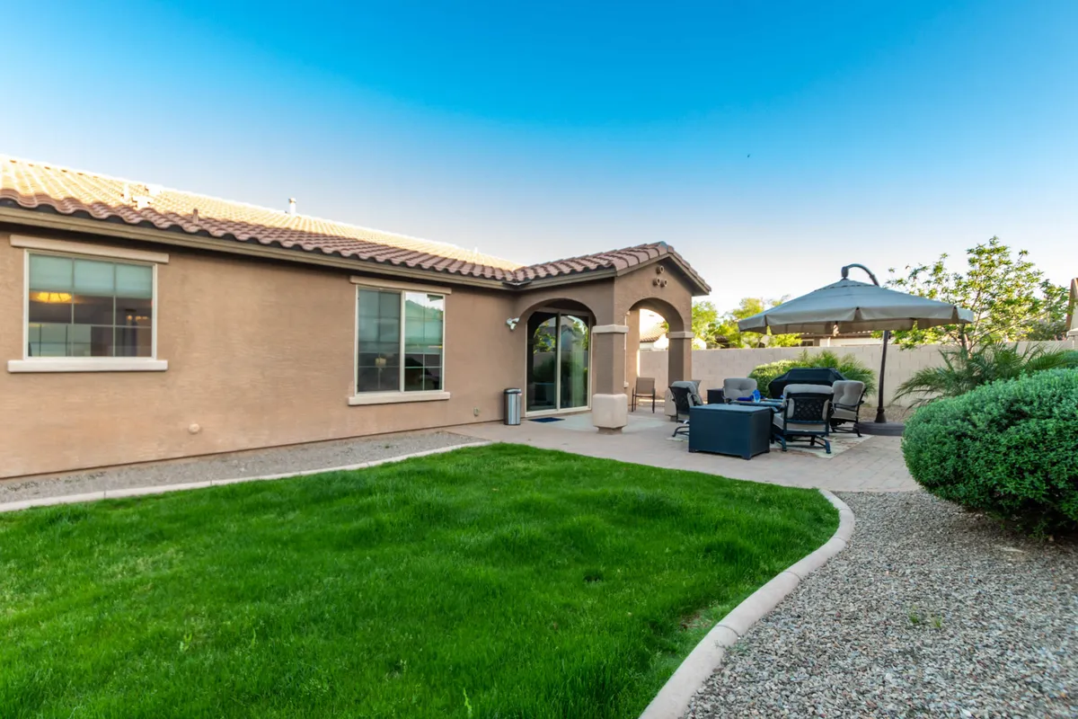 A tranquil backyard scene features a lush, green lawn and comfortable patio furniture set beneath an arched entryway. The peaceful garden is perfect for relaxing in the shade, hosting outdoor meals, or supervising children as they play on the grass.