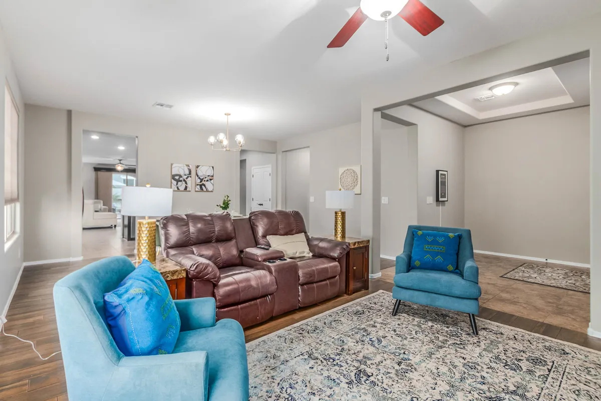 A spacious open-concept living area is anchored by a plush brown leather sectional sofa and accented by blue chairs and a decorative rug. The arrangement provides ample seating for group relaxation, game nights, or movie evenings, and the chandelier overhead adds a touch of warmth and elegance.