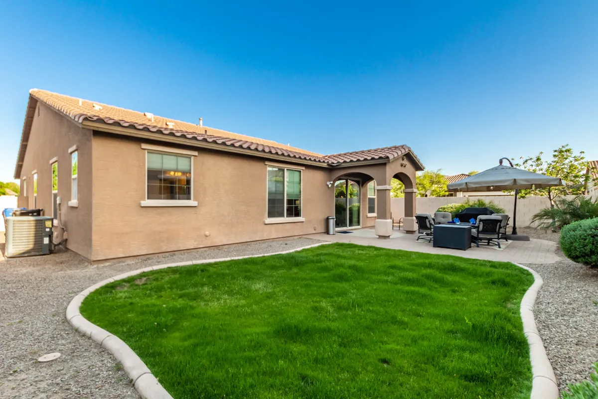 A spacious, lush green lawn stretches across the foreground, bordered by a pergola and shaded patio seating area. Architectural arches offer a welcoming, family-friendly touch, and the tranquil space is perfect for gatherings, games, or relaxed afternoons. The area is surrounded by vibrant landscaping, providing a safe and open environment for children to play under supervision.
