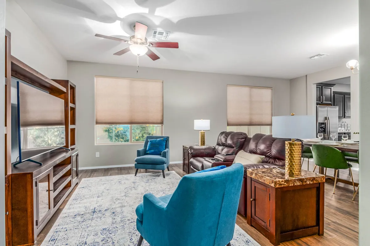 A cozy living area with a brown leather sofa, blue accent chair, a stylish wooden entertainment unit, and a decorative coffee table. The room is arranged to offer both relaxation and entertainment, with the TV as a focal point and natural light filtering in from the windows.