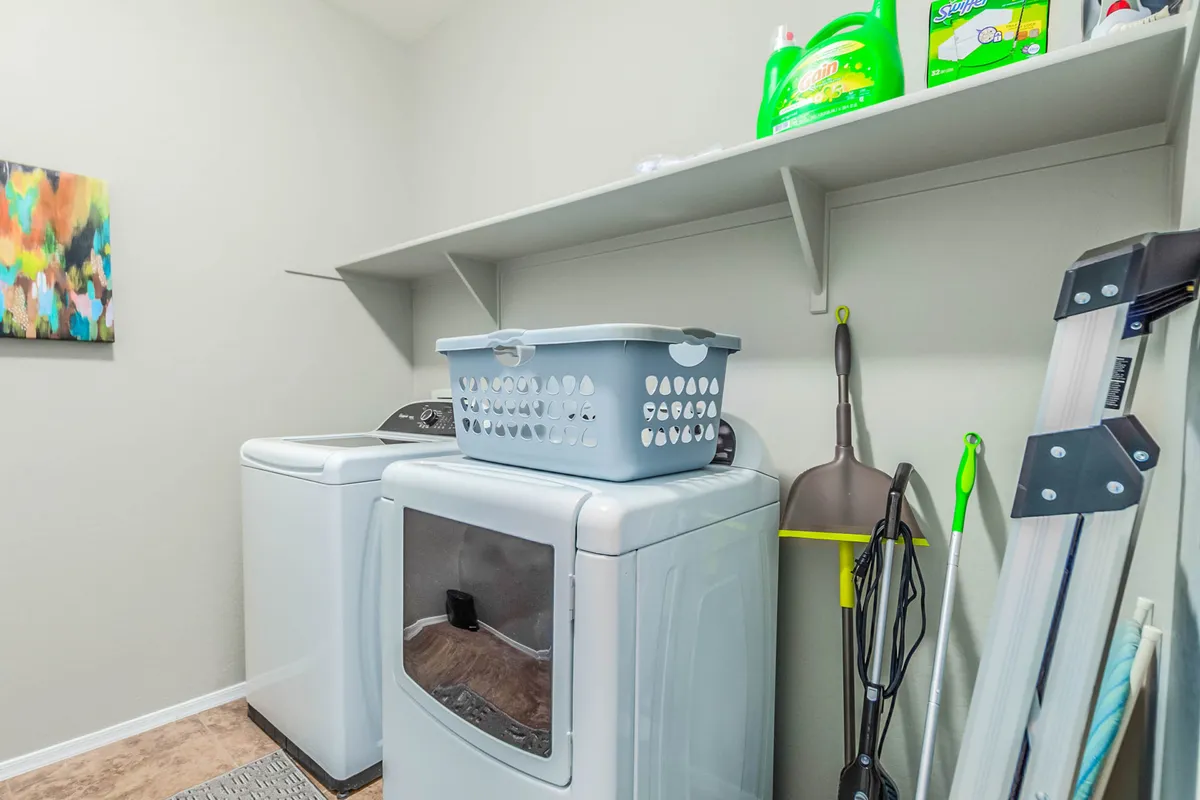 A tidy and practical laundry room, complete with a washer and dryer, a laundry basket, and organized cleaning supplies on shelving. The scene is bright and functional, supporting efficient day-to-day routines.